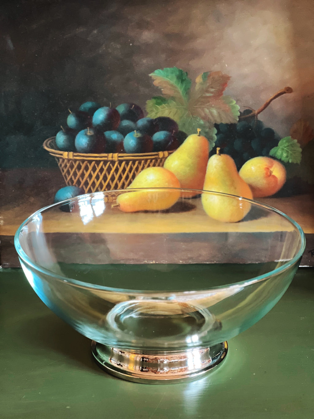 Clear glass bowl on a dark surface with a still life painting of fruit in the background