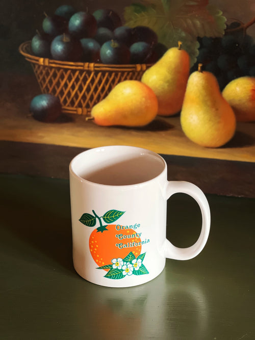 White mug with 'Orange County, California' design on a reflective surface with fruit in the background