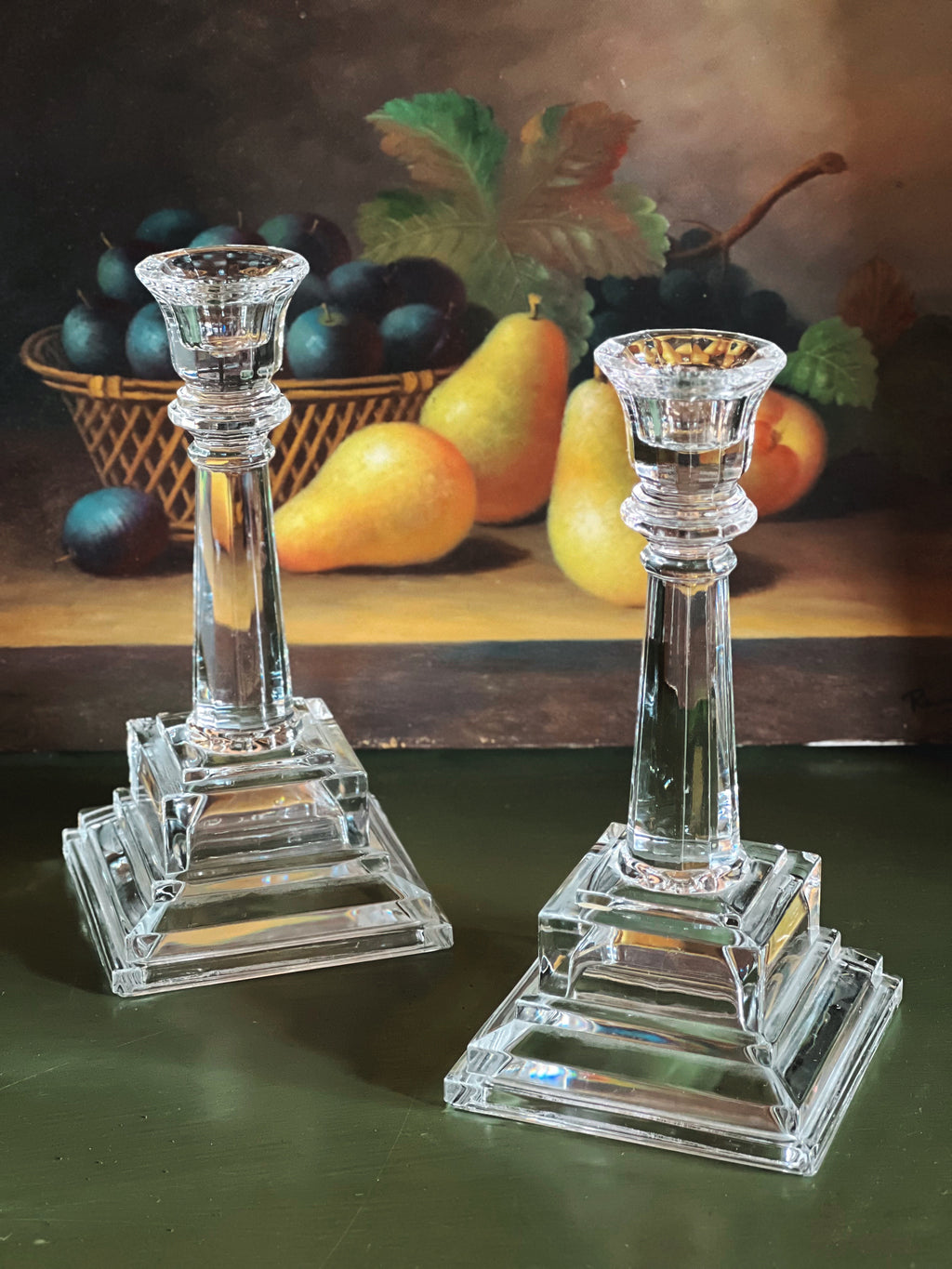 Two crystal candlesticks on a dark surface with a still life painting of fruit in the background.