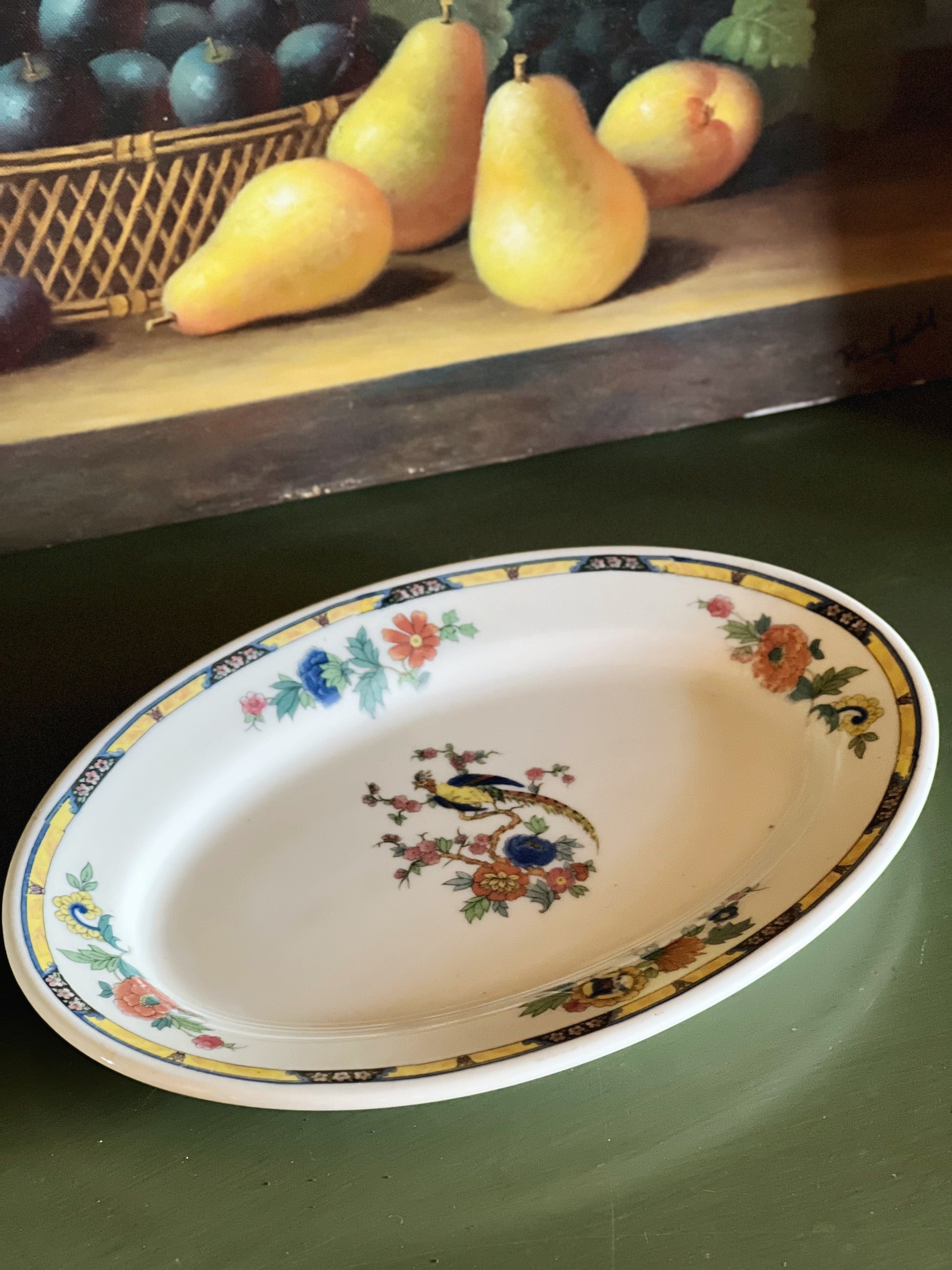Decorative oval plate with floral and bird design on a green surface, with a painting of pears and apples in the background.