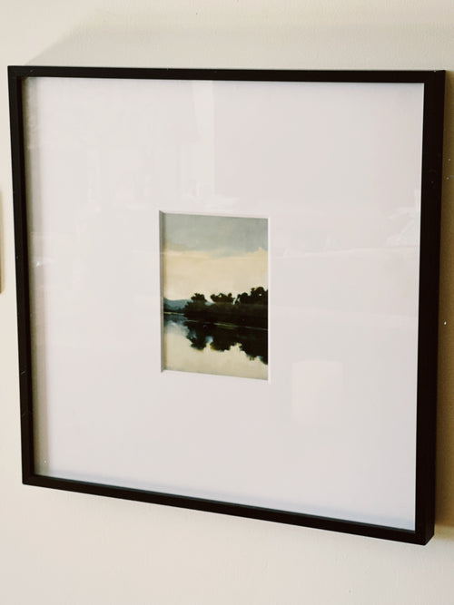Framed photograph of a landscape with trees and water on a white wall.