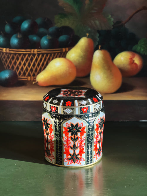 Decorative jar with floral patterns on a reflective surface with fruits in the background