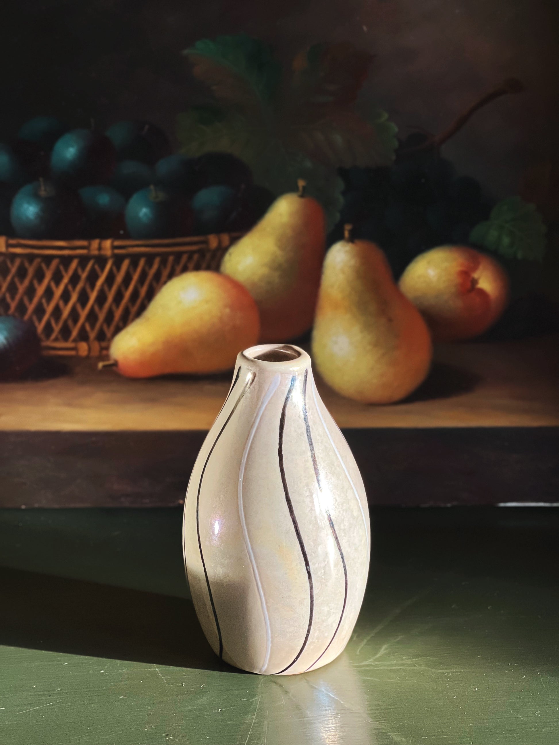 White vase with black stripes on a dark surface with pears and a basket in the background