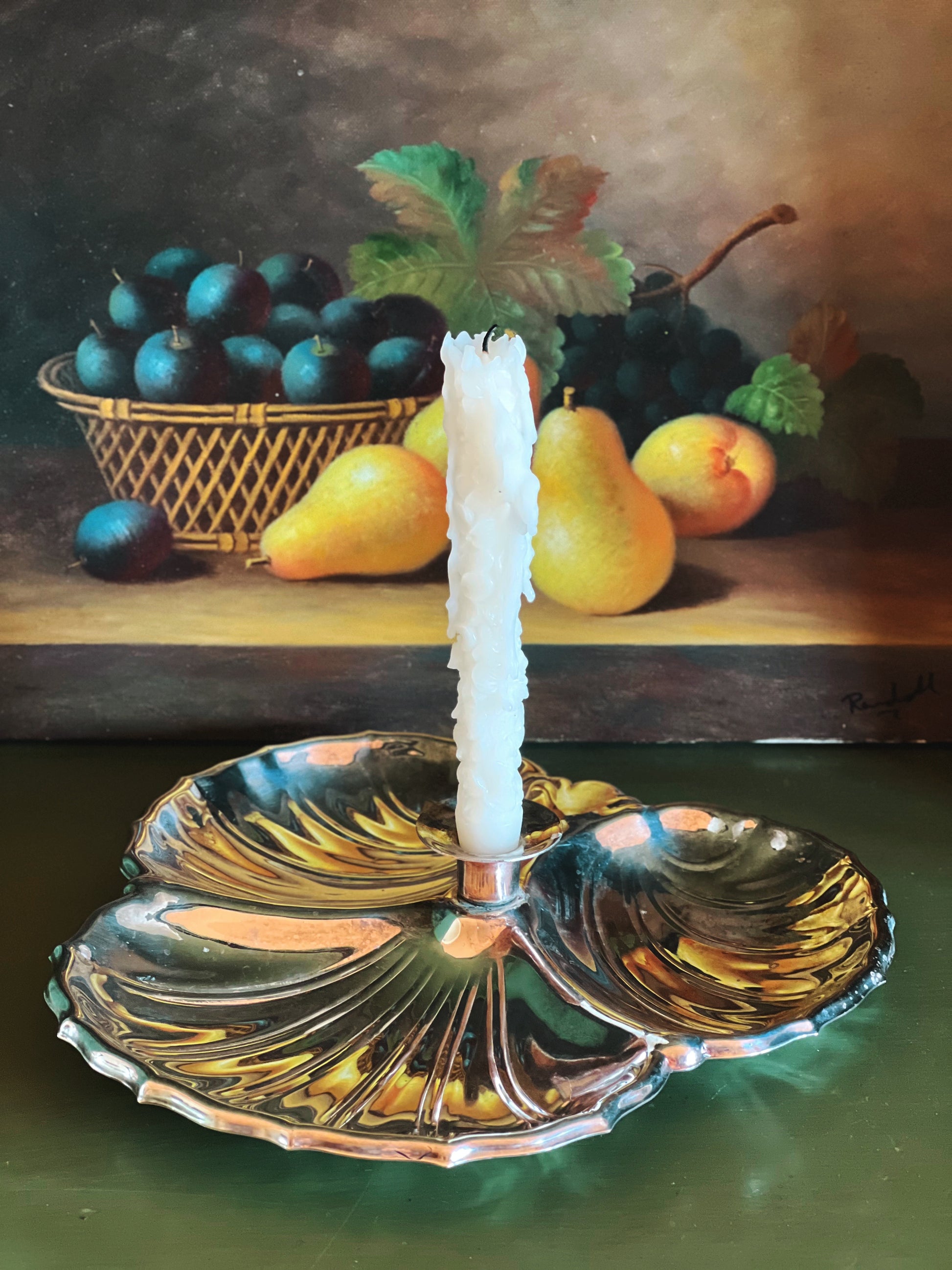 Still life painting of fruits and a candle with a decorative silver dish in the foreground.