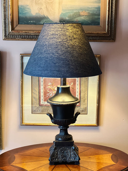 Decorative lamp with a black shade on a wooden table against a wall with framed pictures.