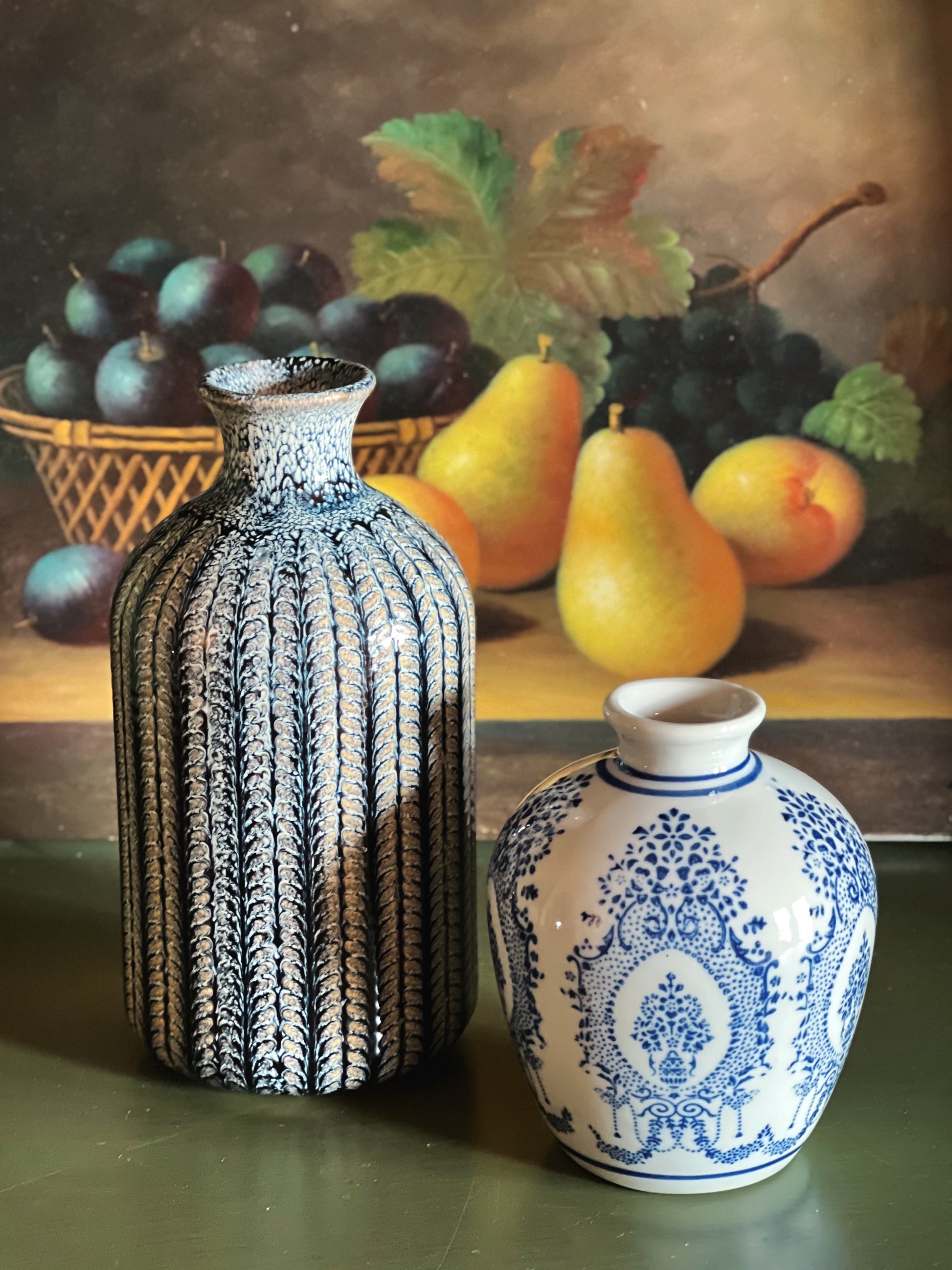 Two decorative vases on a table with a still life painting of fruit in the background.