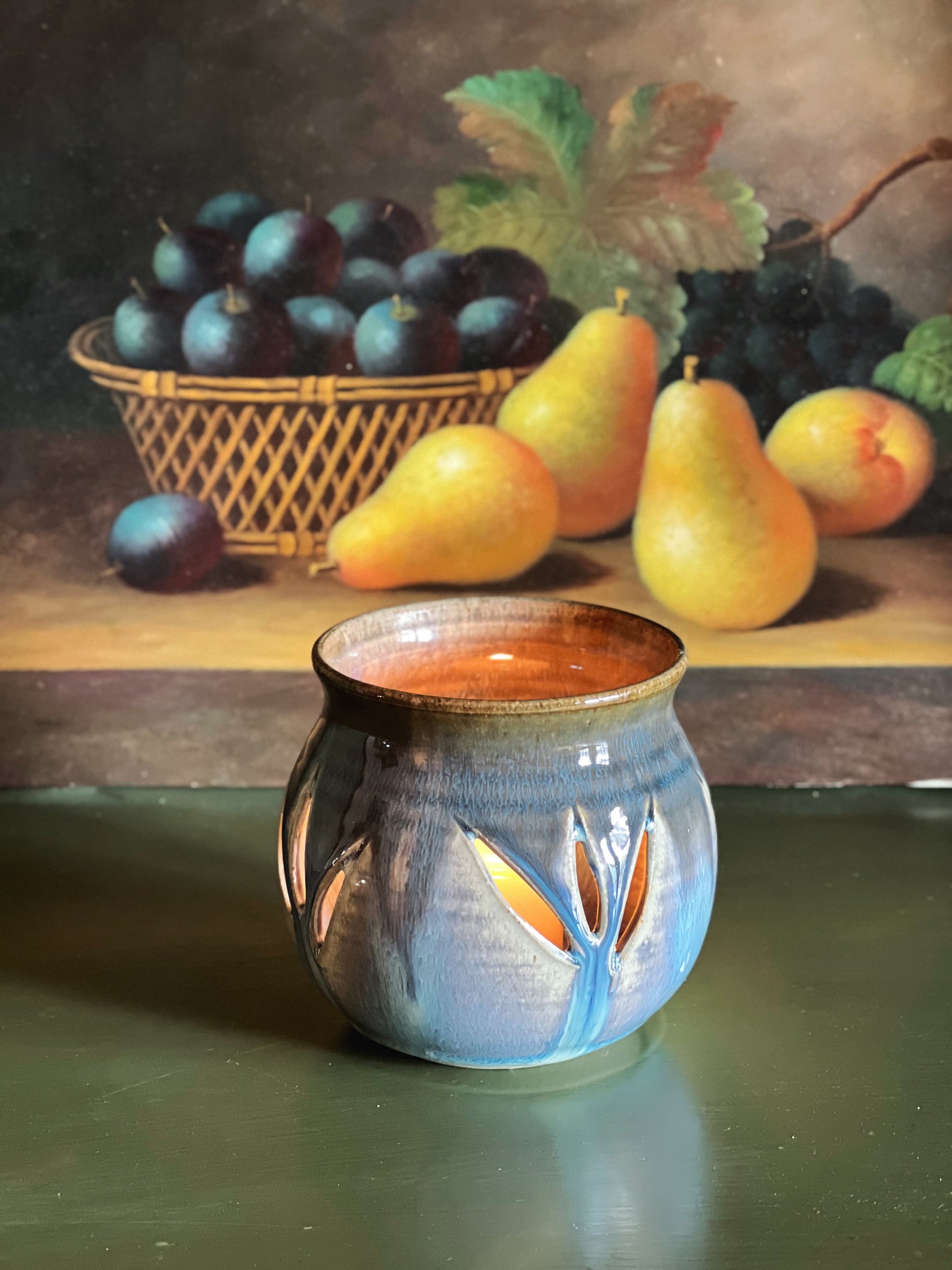 Ceramic pot with floral design on a reflective surface, with a still life painting of fruit in the background.