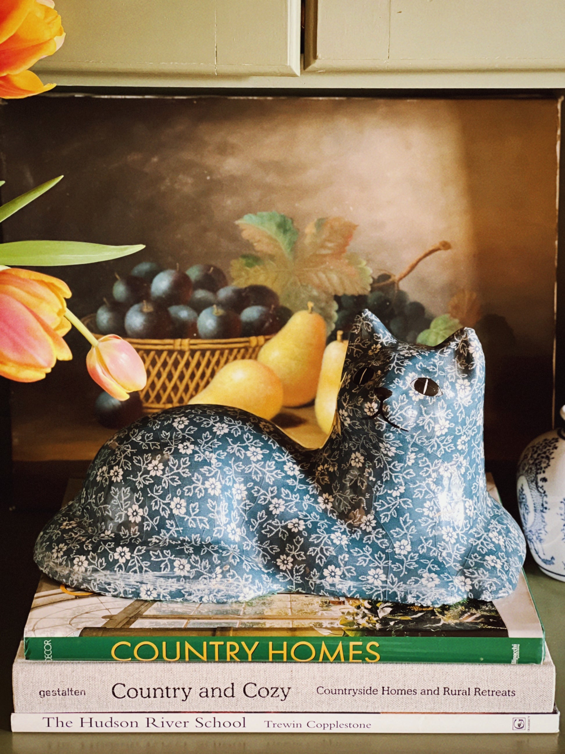 Decorative blue bird figurine on a book titled 'Country Homes' with tulips and a painting in the background.