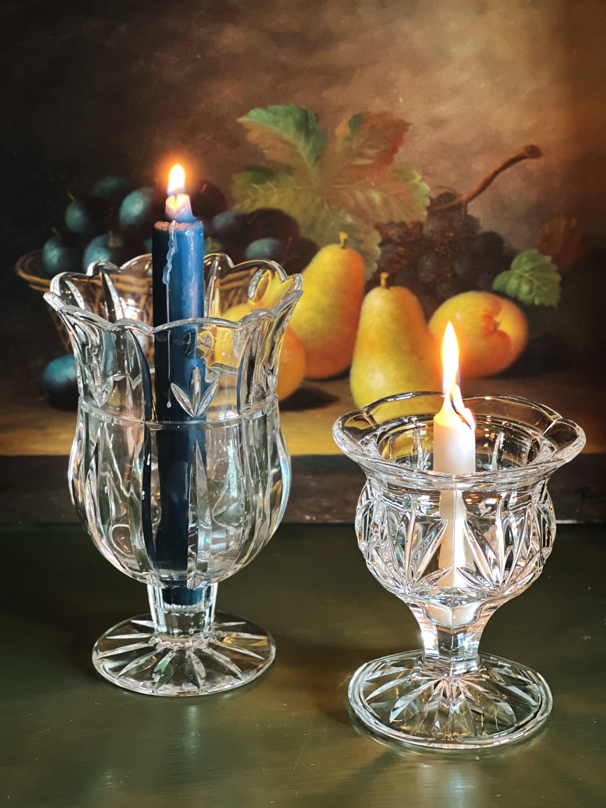 Two glass candle holders with lit candles on a table with fruit in the background.