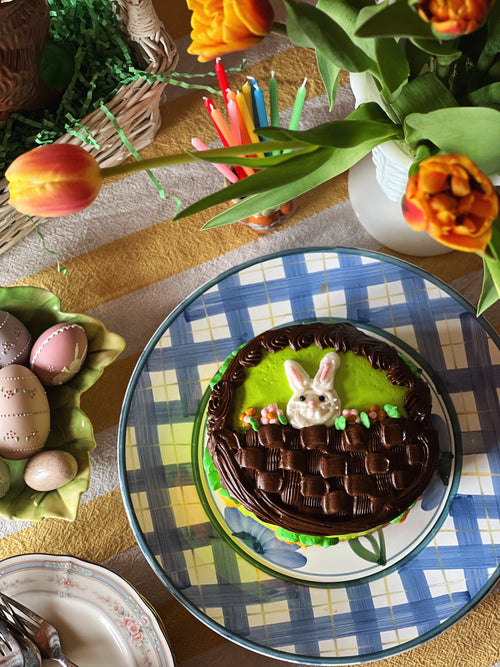 Chocolate Easter cake with bunny decoration on a checkered tablecloth, surrounded by tulips and Easter eggs.