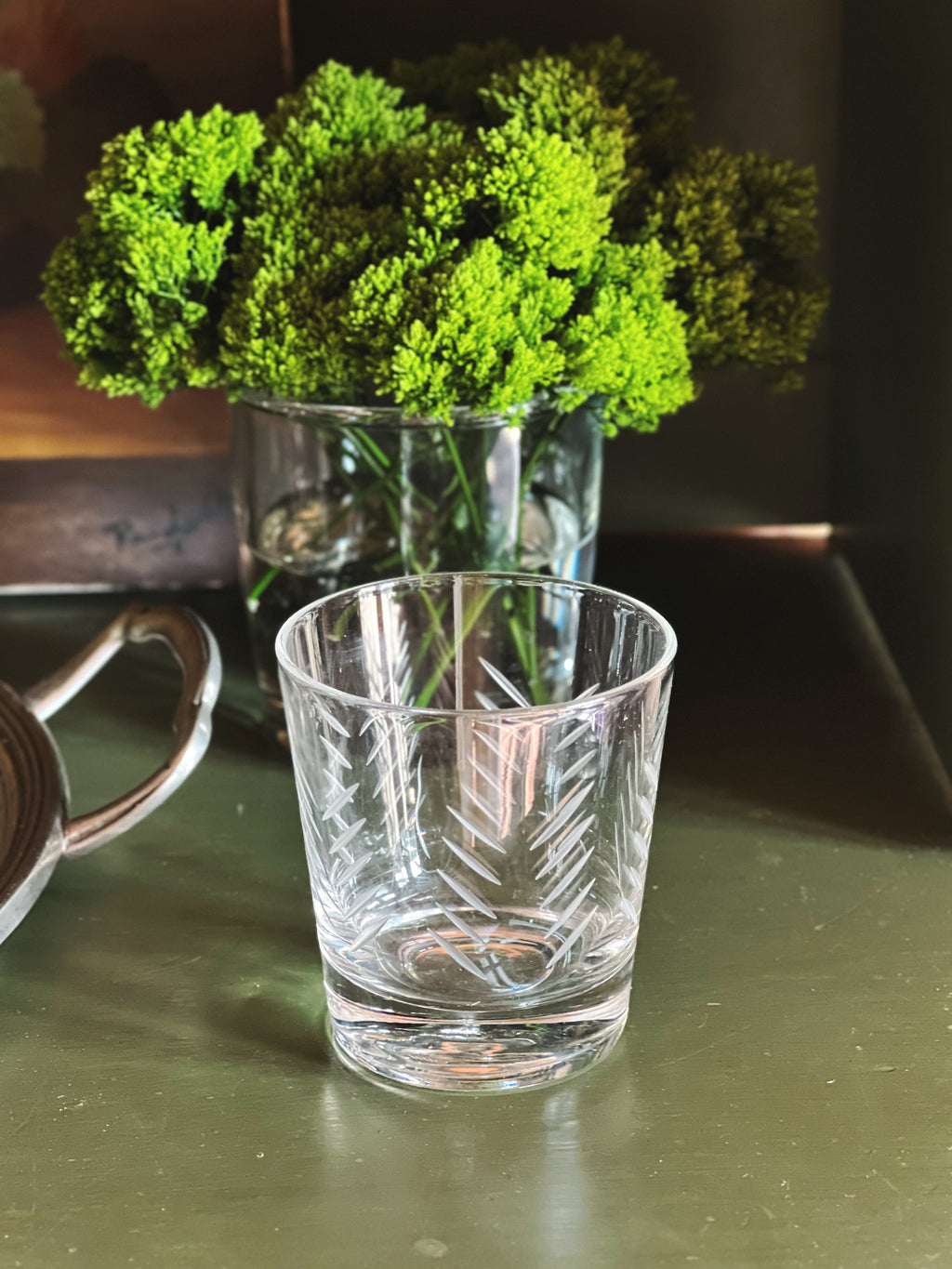 Clear glass with engraved design on a reflective surface, next to a vase of green flowers.