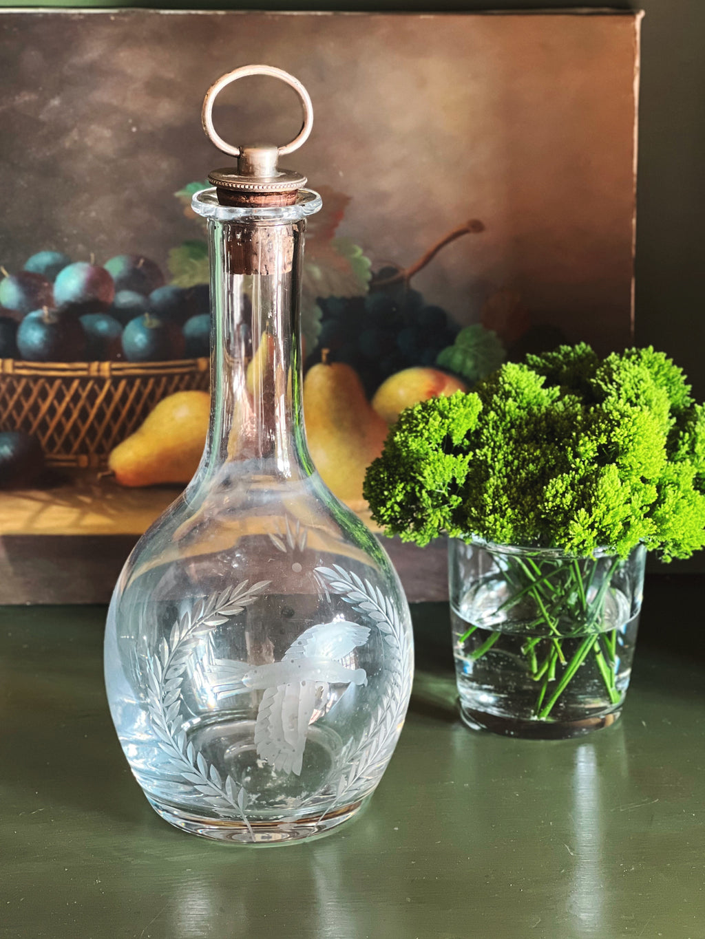 Clear glass Wedgwood decanter with engraved design on a table with fruit and greenery in the background.