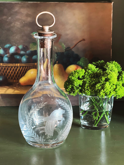 Clear glass Wedgwood decanter with engraved design on a table with fruit and greenery in the background.