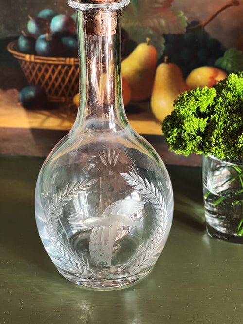 Clear glass Wedgwood decanter with engraved bird and laurel design on a wooden surface with fruits and vegetables in the background