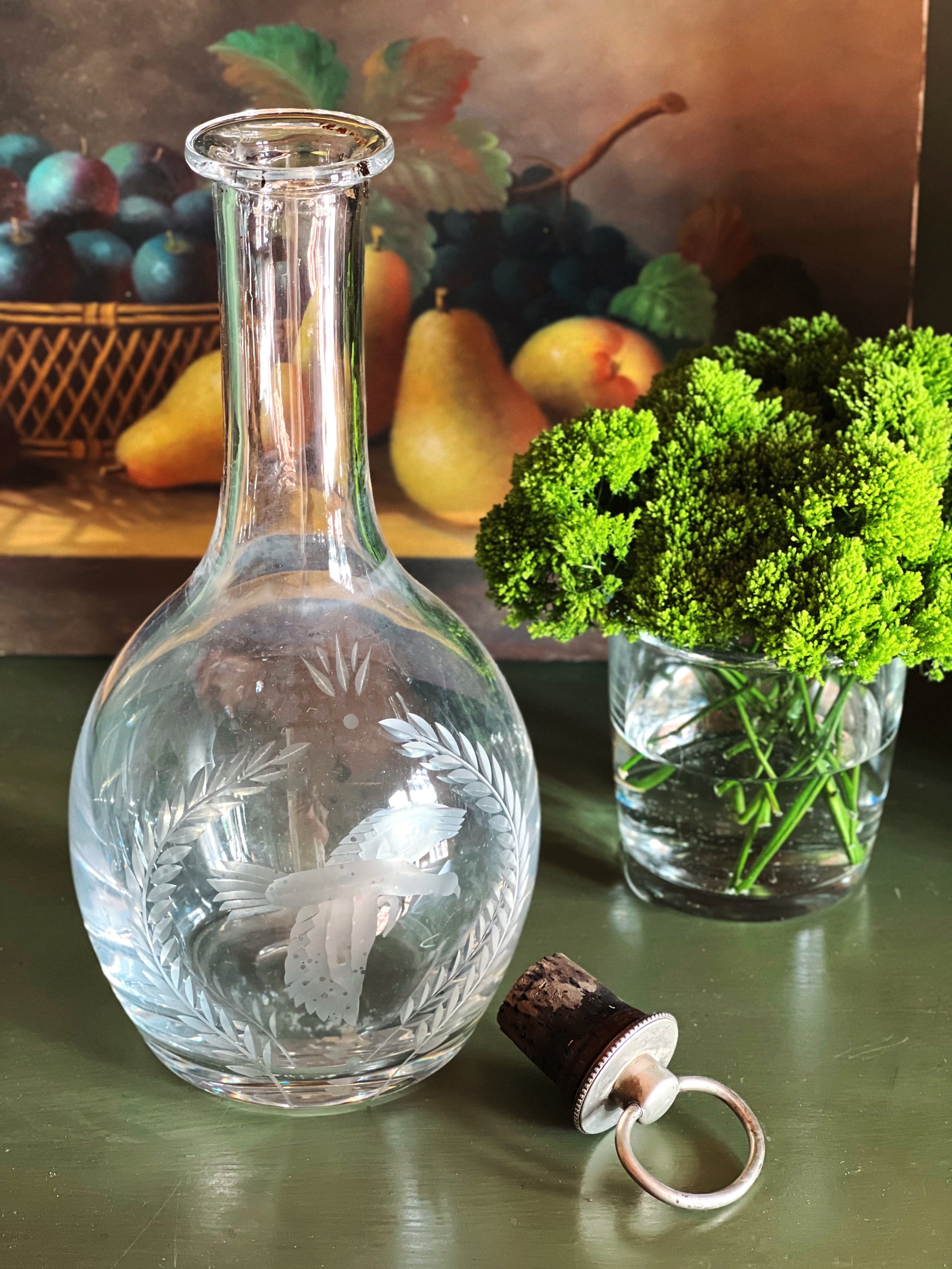 Wedgwood glass decanter with engraved design and cork on a table with a painting of fruit in the background.