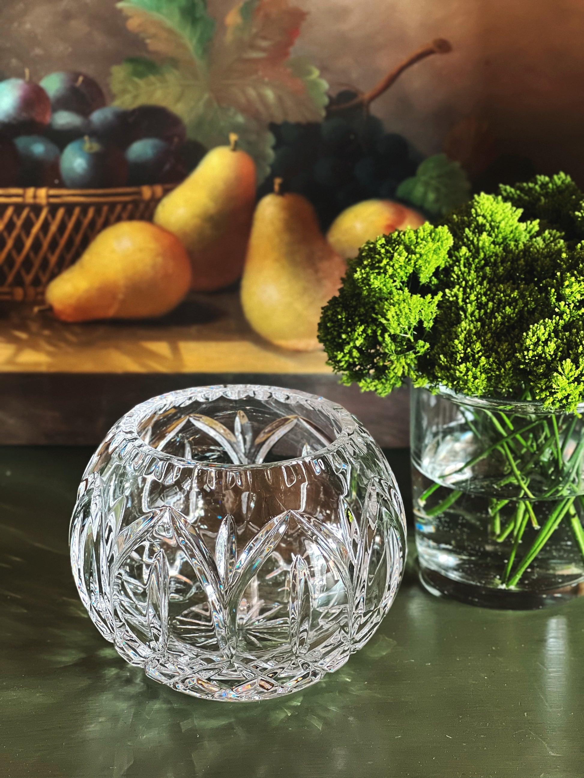 Side angle of a crystal bowl on a reflective surface with a fruit painting in the background