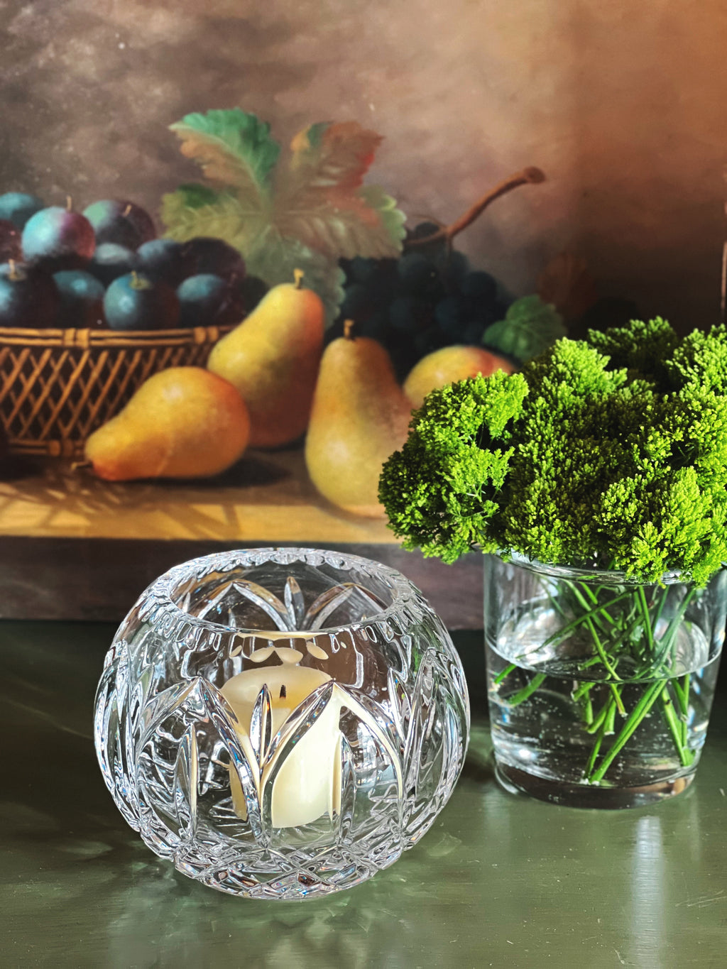 Decorative crystal candle holder with a lit candle, surrounded by pears, grapes, and a vase of greenery on a wooden surface.
