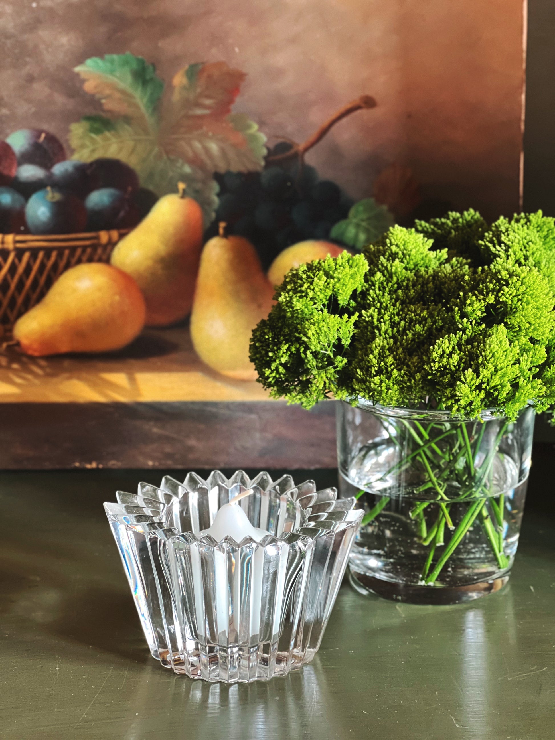Clear glass votive and vase with greenery on a reflective surface, with a painting of fruit in the background.