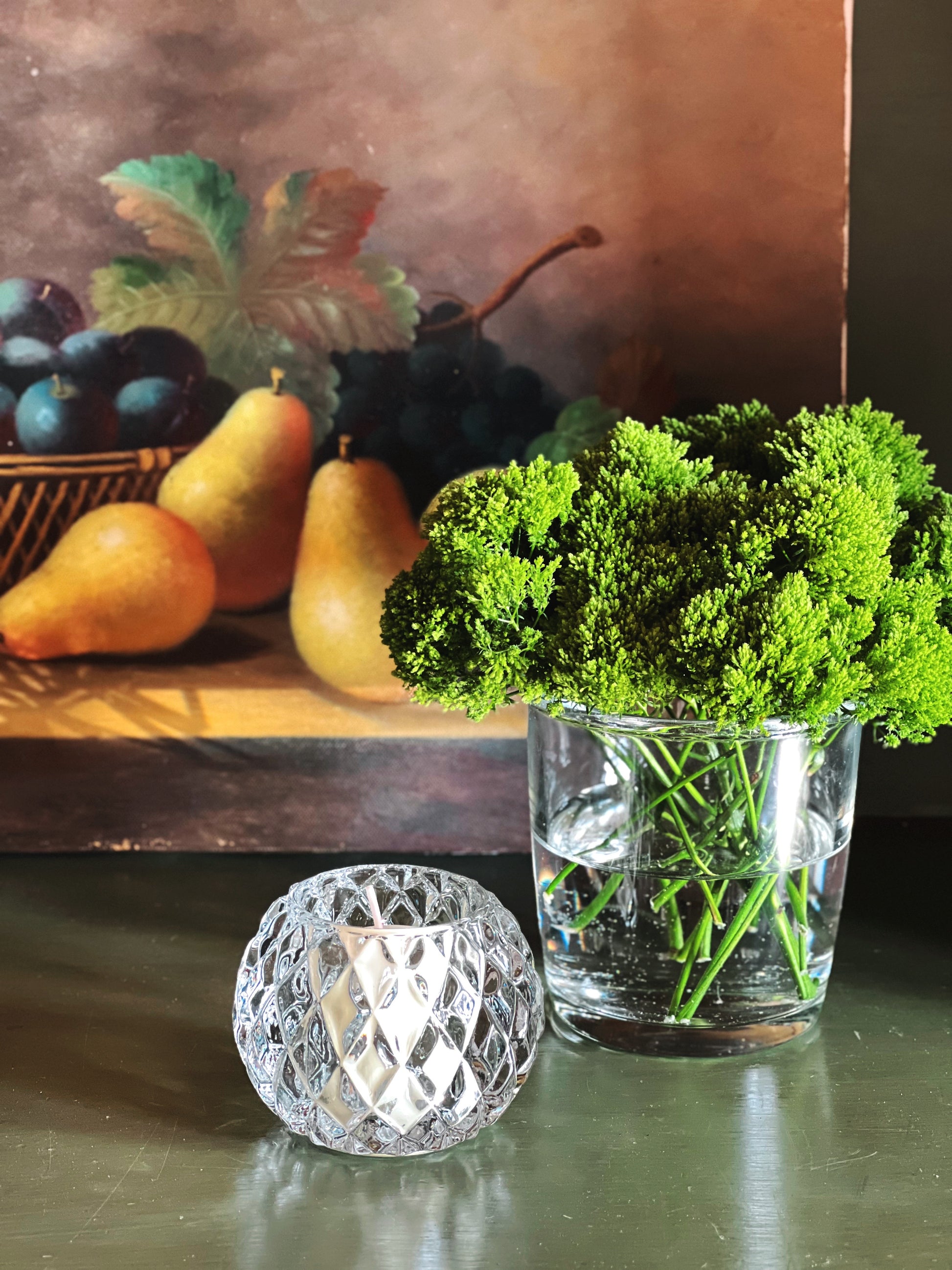 Still life arrangement with pears, a basket of plums, a glass vase with greenery, and a glass votive holder on a reflective surface.