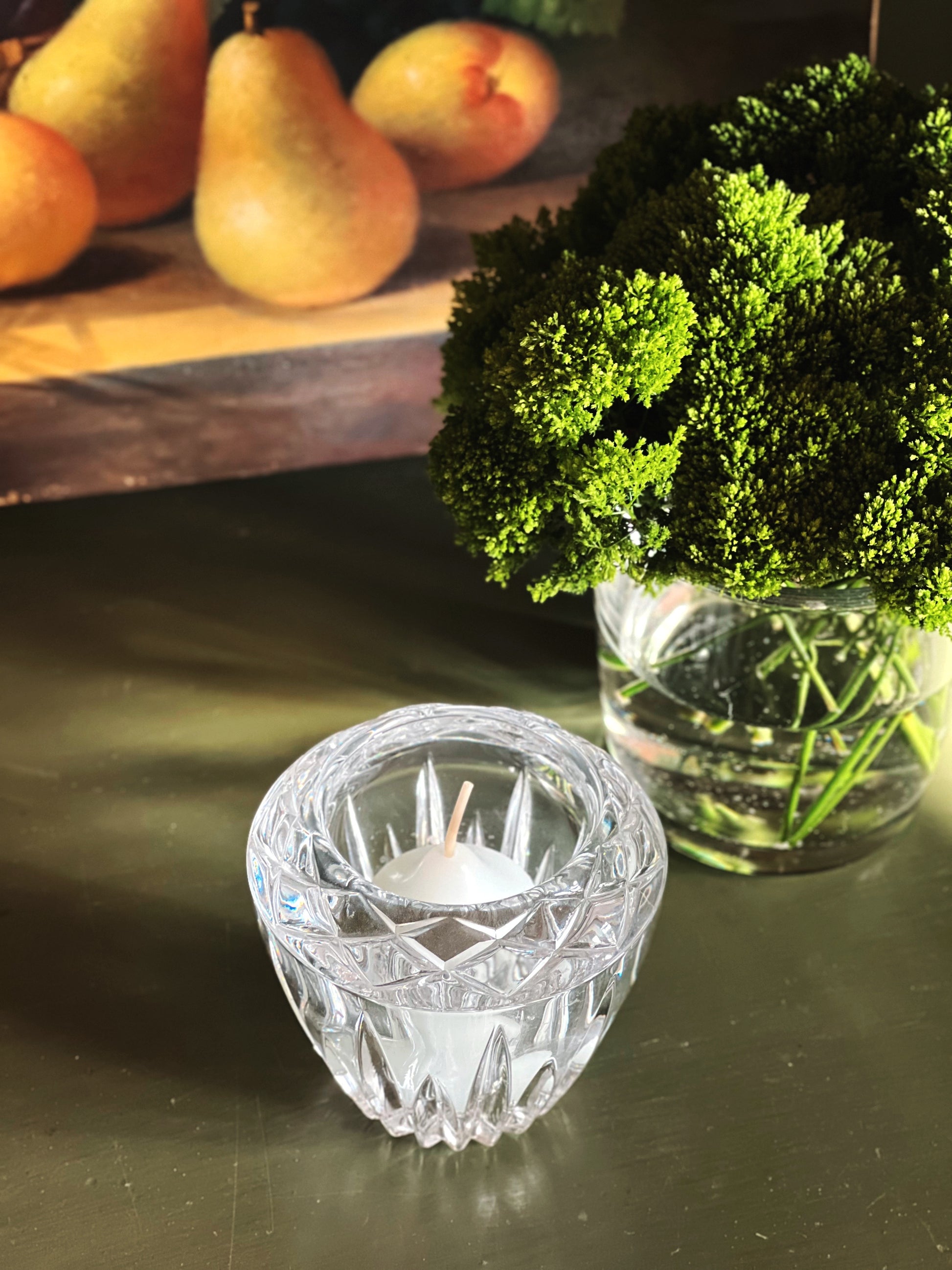 Crystal candle holder with a small candle on a reflective surface, with pears and a plant in the background.