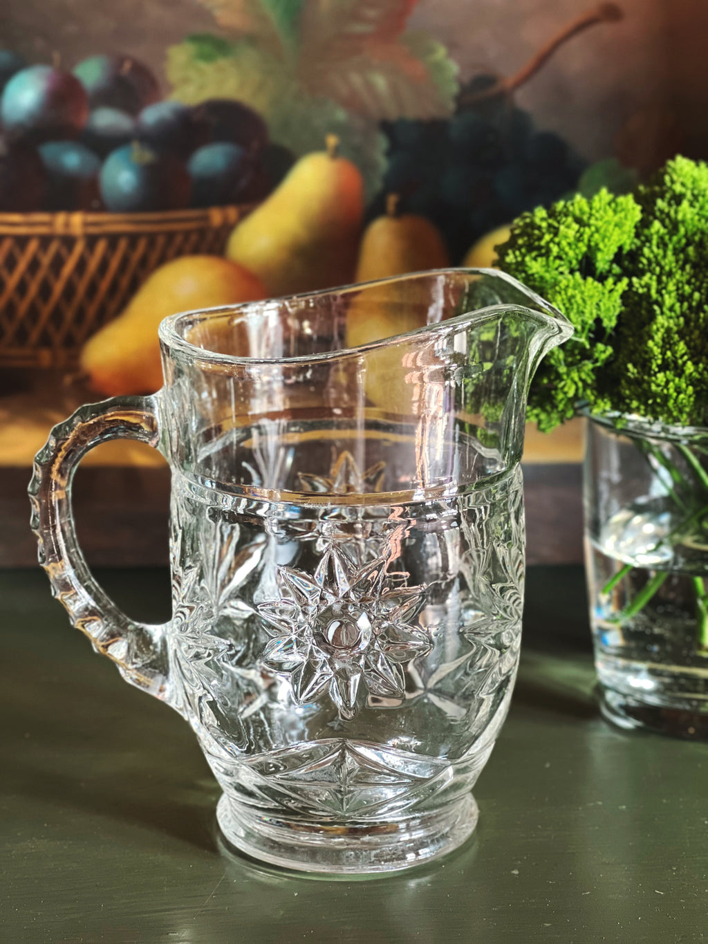 Vintage cut glass cream pitcher with starburst pattern styled on a dark green surface beside fruit and greenery