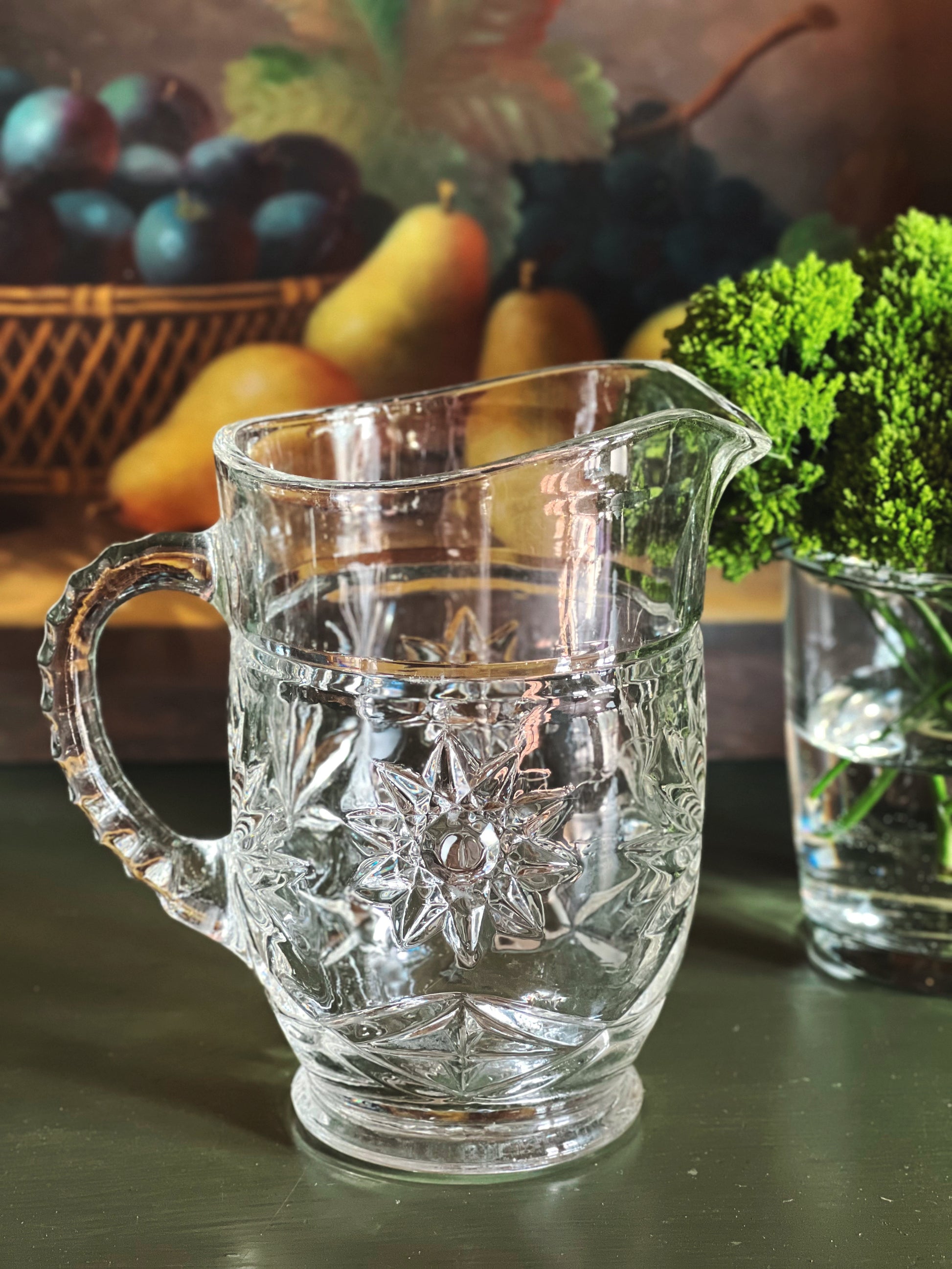 Vintage cut glass cream pitcher with starburst pattern styled on a dark green surface beside fruit and greenery