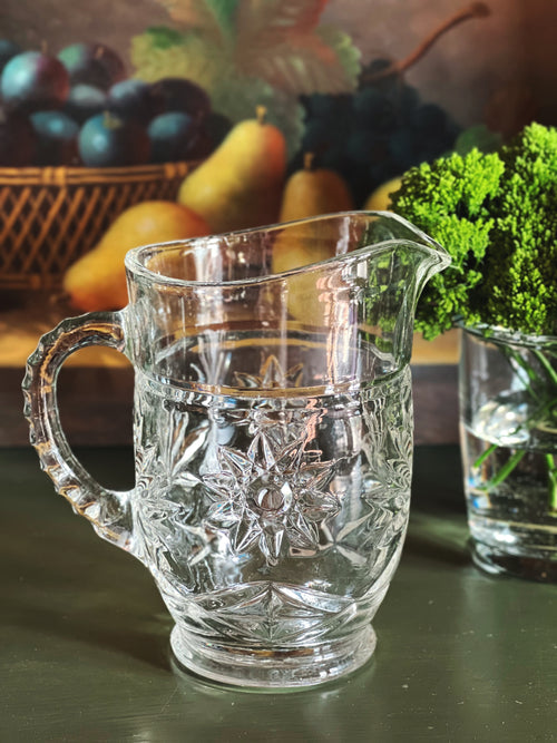 Vintage cut glass cream pitcher with starburst pattern styled on a dark green surface beside fruit and greenery