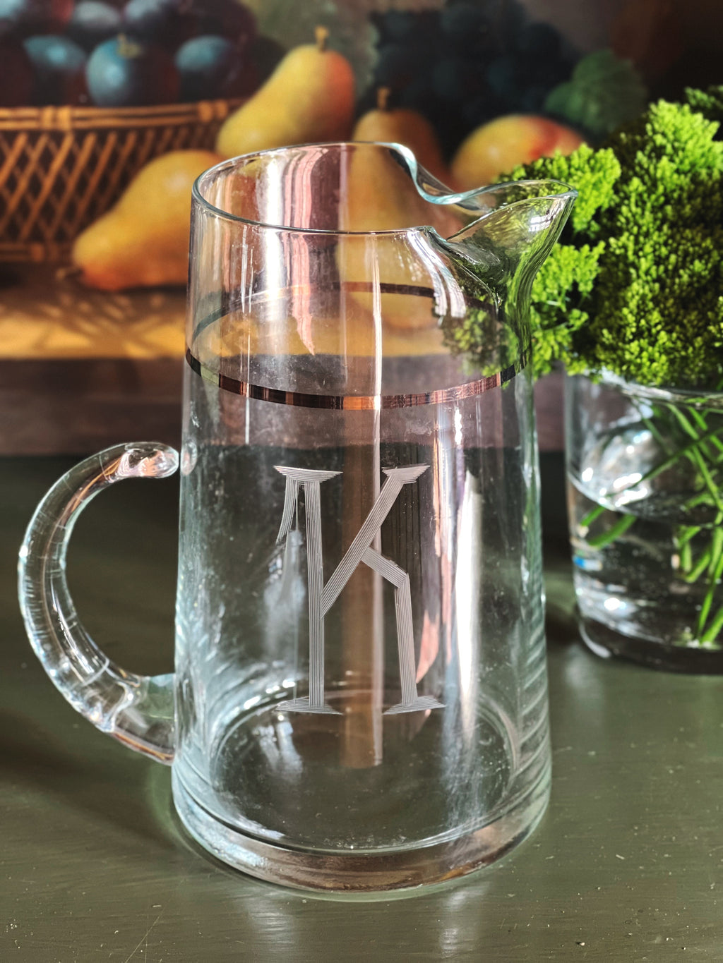 Vintage monogram glass pitcher with silver band detail and sculptural spout styled on a dark green surface