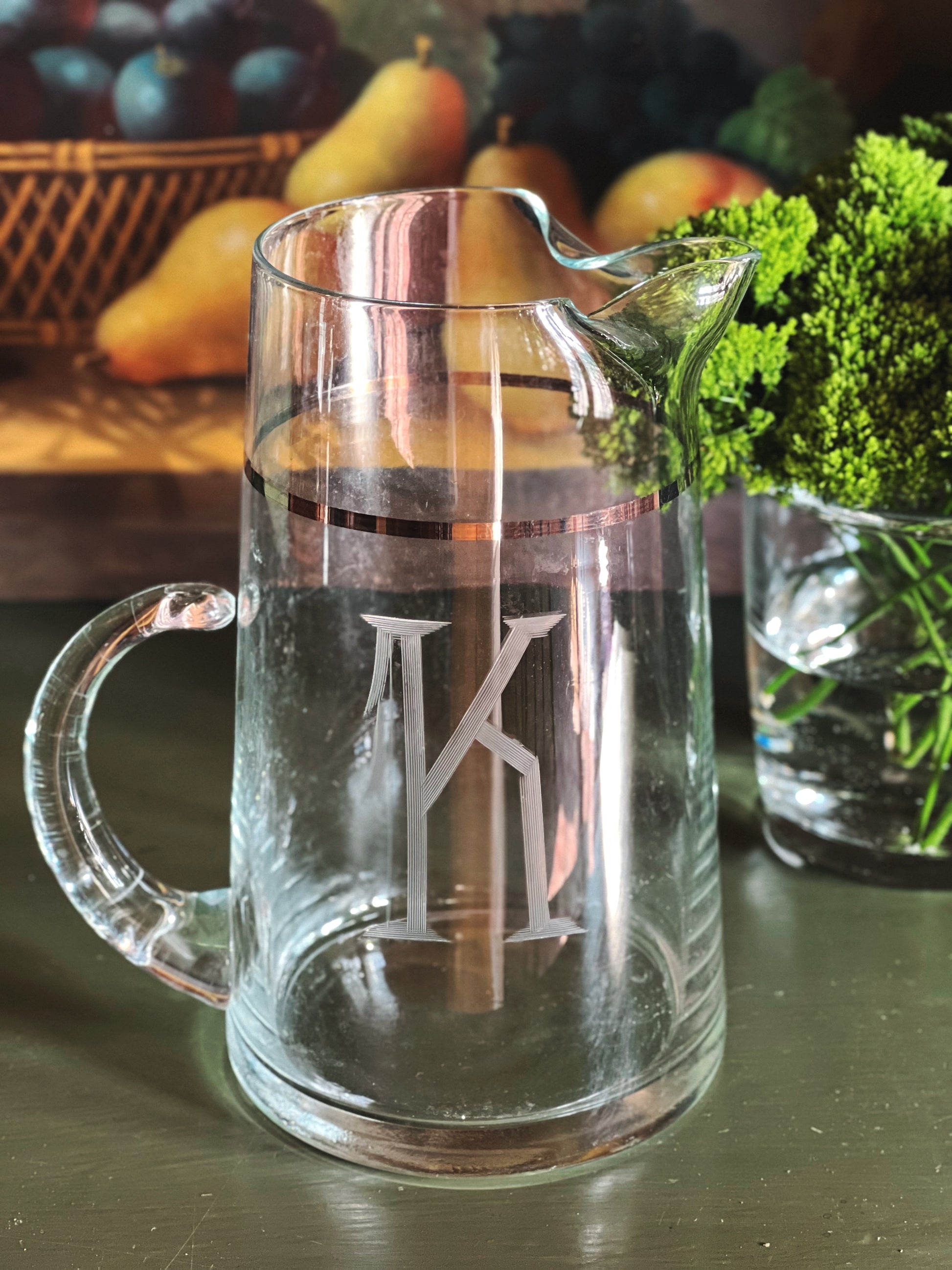 Vintage monogram glass pitcher with silver band detail and sculptural spout styled on a dark green surface