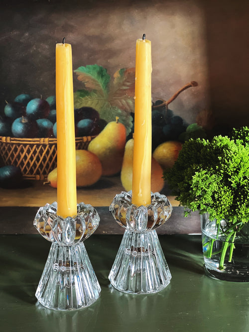 Two yellow candles in crystal candle holders on a table with a fruit basket and plant in the background.
