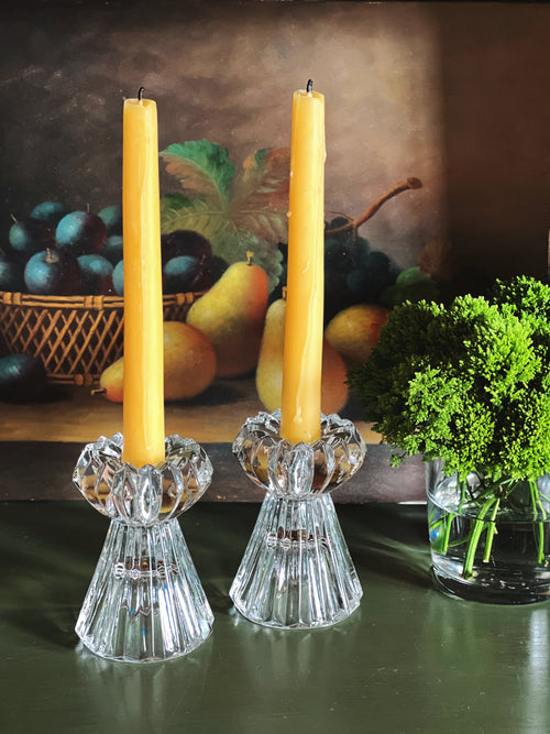Two beeswaxcandles in crystal candle holders on a table with a fruit basket and plants in the background.