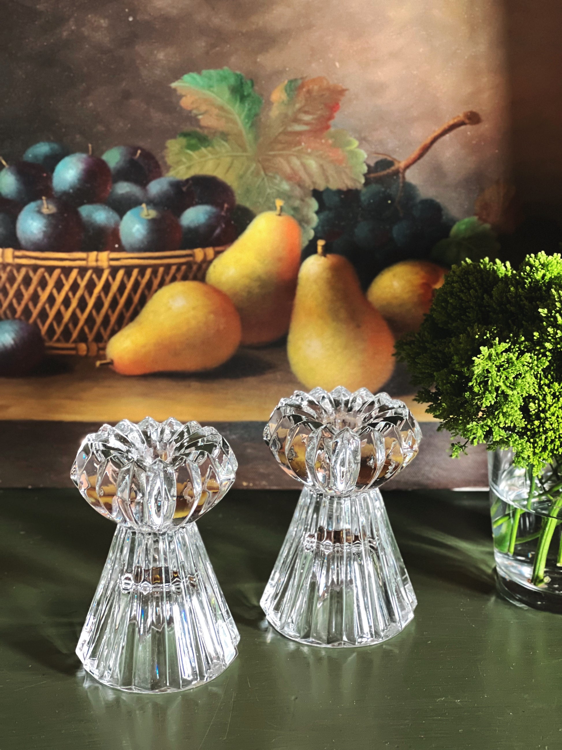 Two crystal candle holders on a table with a fruit basket and painting in the background.