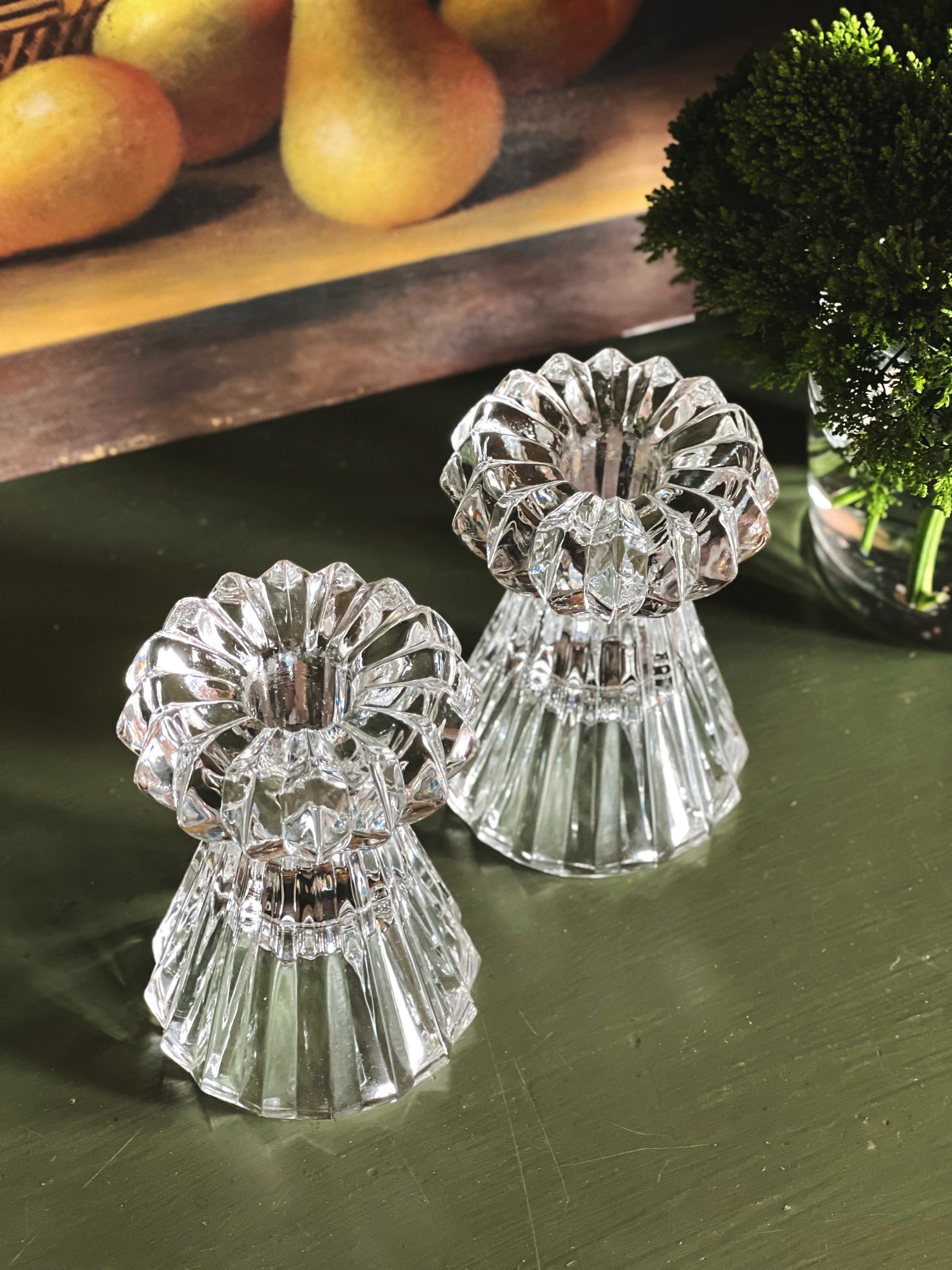 Overhead view of two crystal candle holders on a green surface with fruits and a plant in the background.