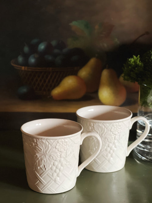 Two Mikasa English Countryside mugs on a wooden surface with fruits and a plant in the background.
