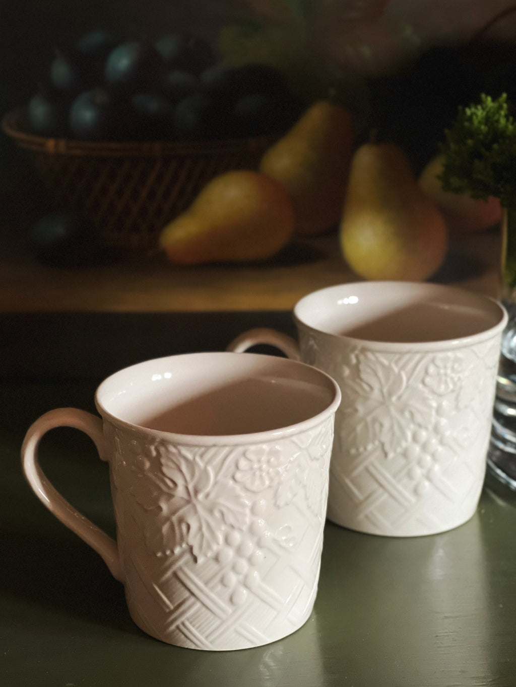 Two white embossed ceramic mugs on a dark surface with a blurred background of fruits and a plant.