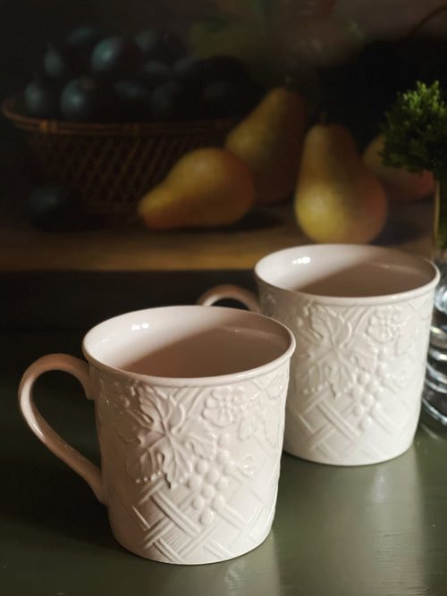 Two white embossed ceramic mugs on a dark surface with a blurred background of fruits and a plant.