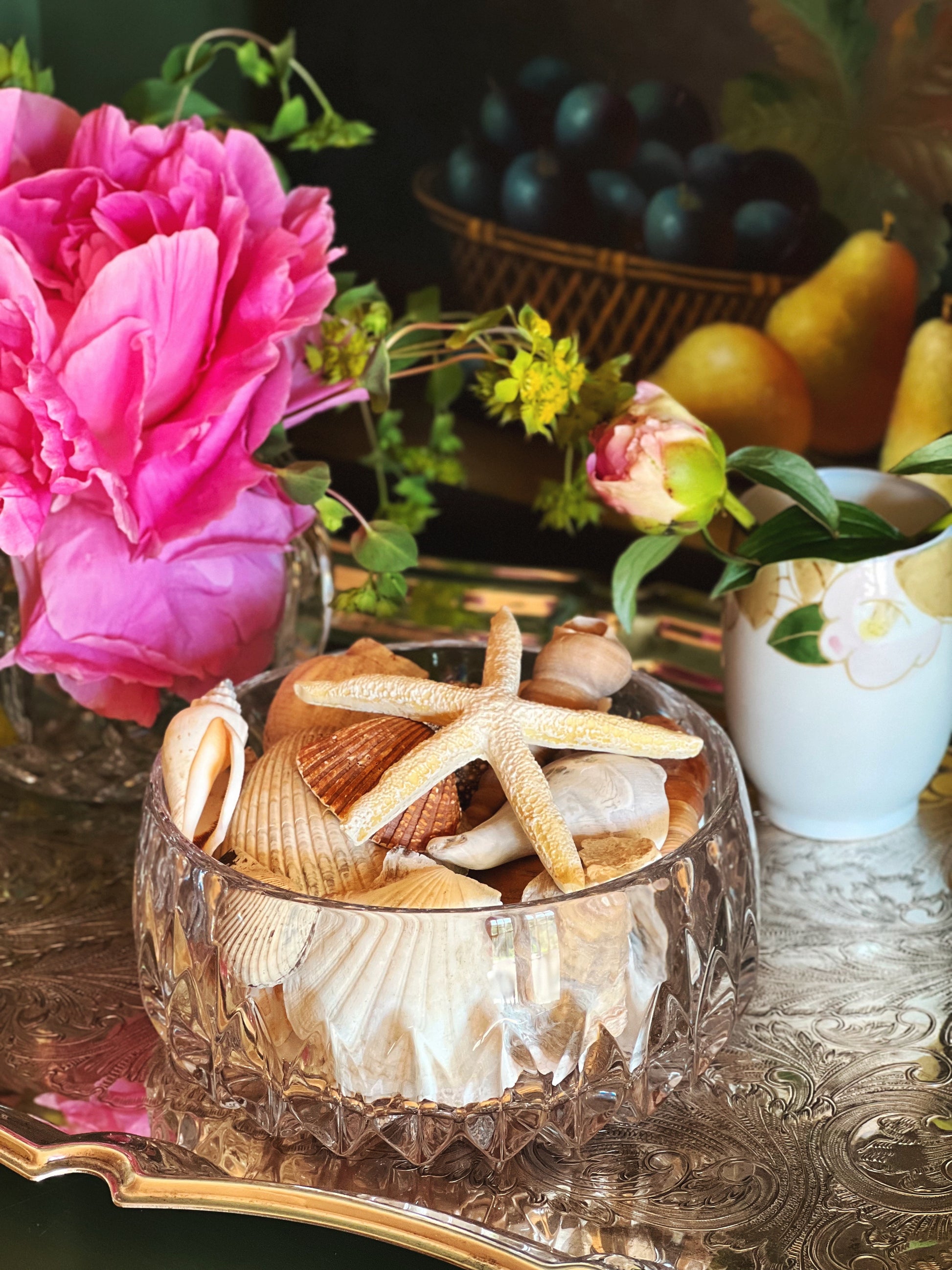 side view of a crystal glass bowl with seashells and starfish on a table with flowers and fruit in the background.