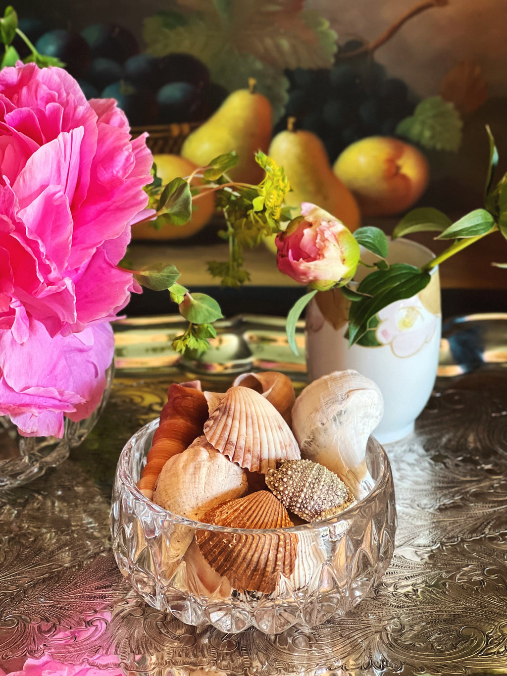 Clear crystal bowl filled with seashells on a decorative table with flowers and fruit in the background.