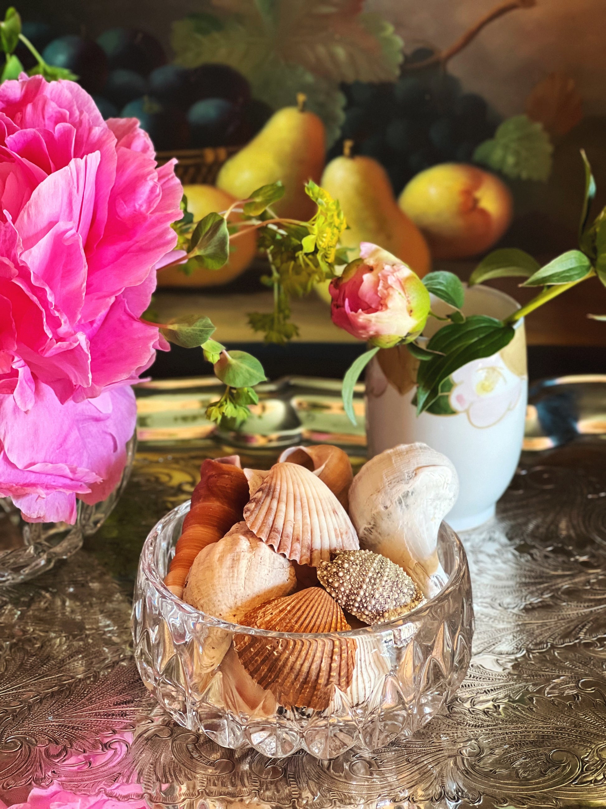 Clear crystal bowl filled with seashells on a decorative table with flowers and fruit in the background.