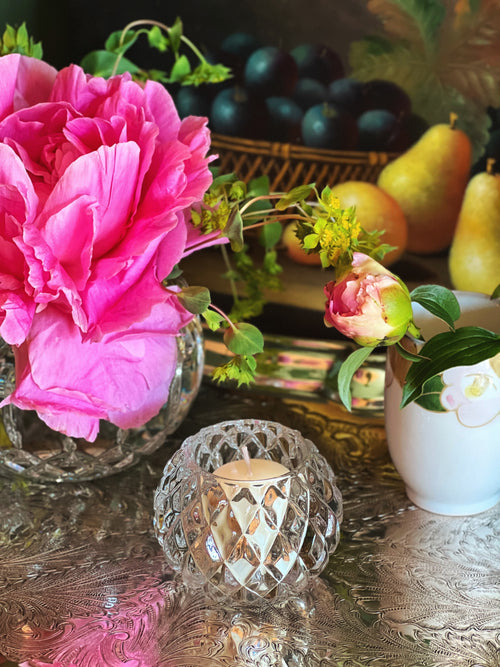 Still life arrangement with pink flowers, fruits, and a bubble glass candle holder on a textured surface.