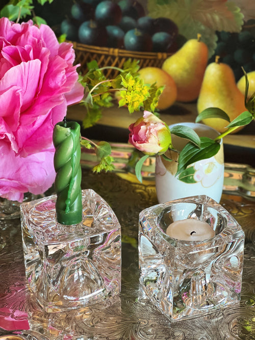 Two reversible crystal candle holders with a pink flower and green candle in the foreground, with fruits and flowers in the background.