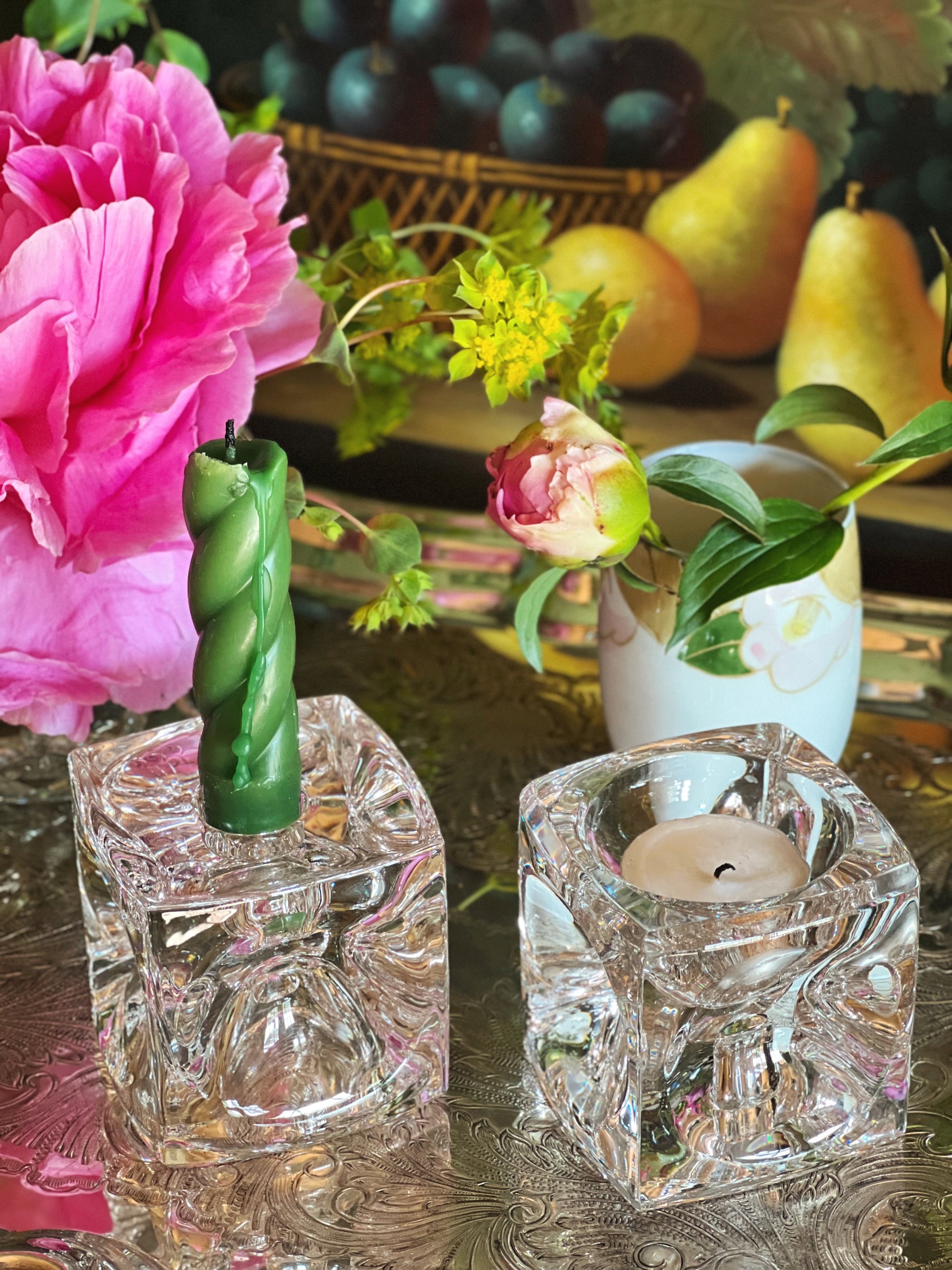 Two crystal candle holders with candles on a table with flowers and fruit in the background.