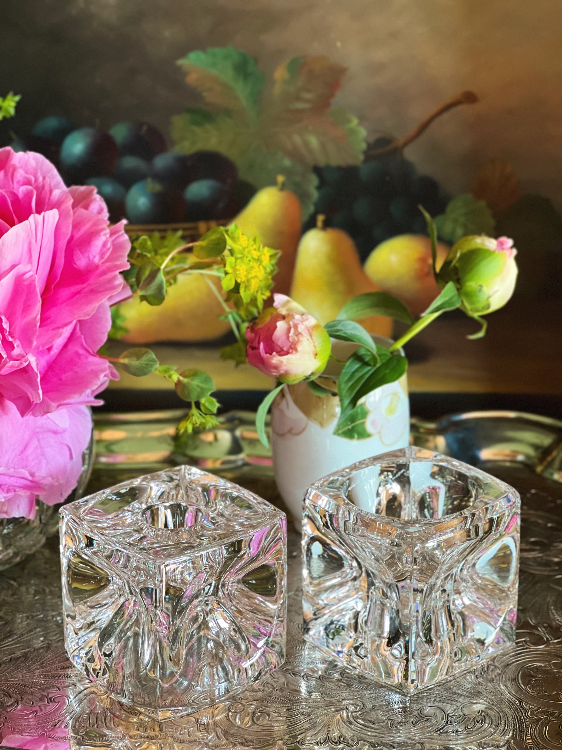 Two crystal candle holders on a reflective surface with flowers and fruit in the background.