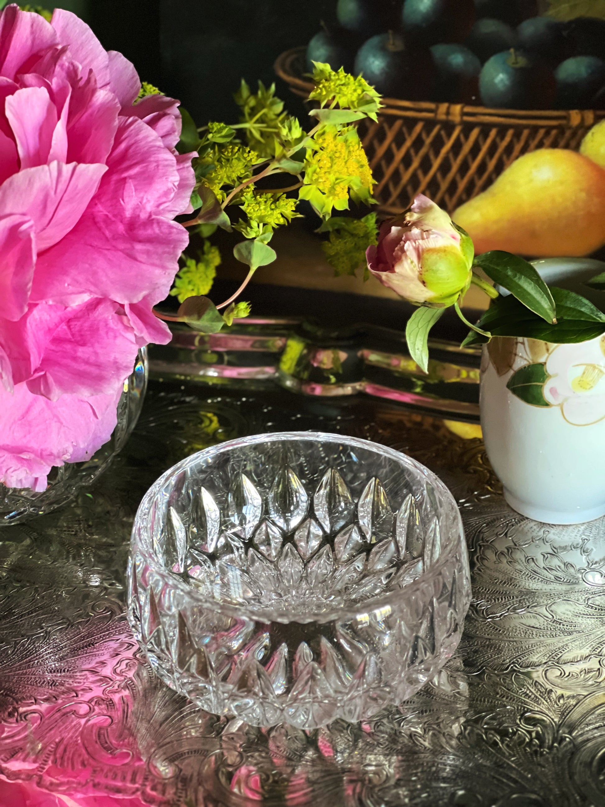 Crystal bowl on a reflective surface with flowers and fruit in the background
