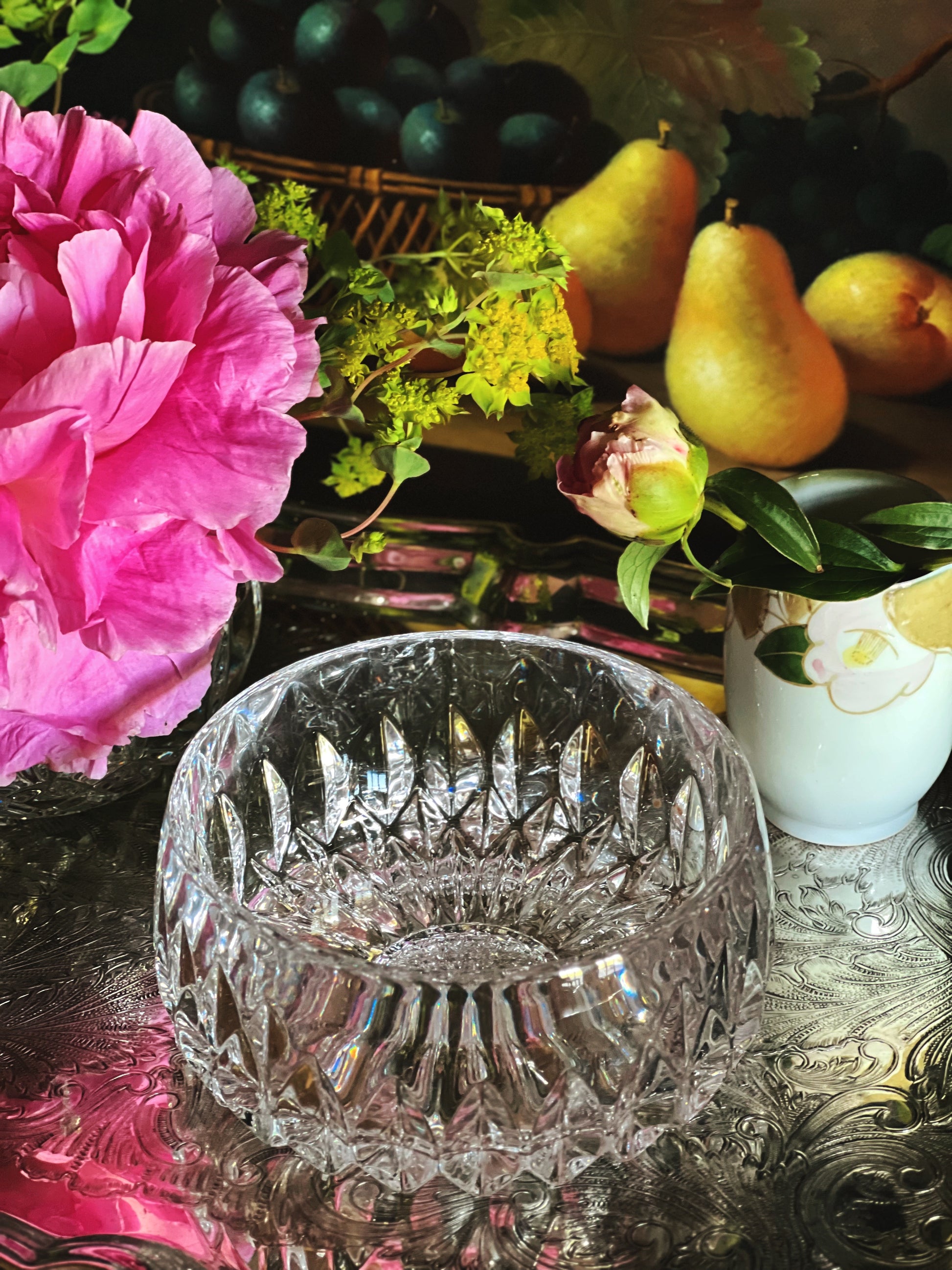 Crystal bowl on a reflective surface with pink flowers, pears, and a basket in the background.