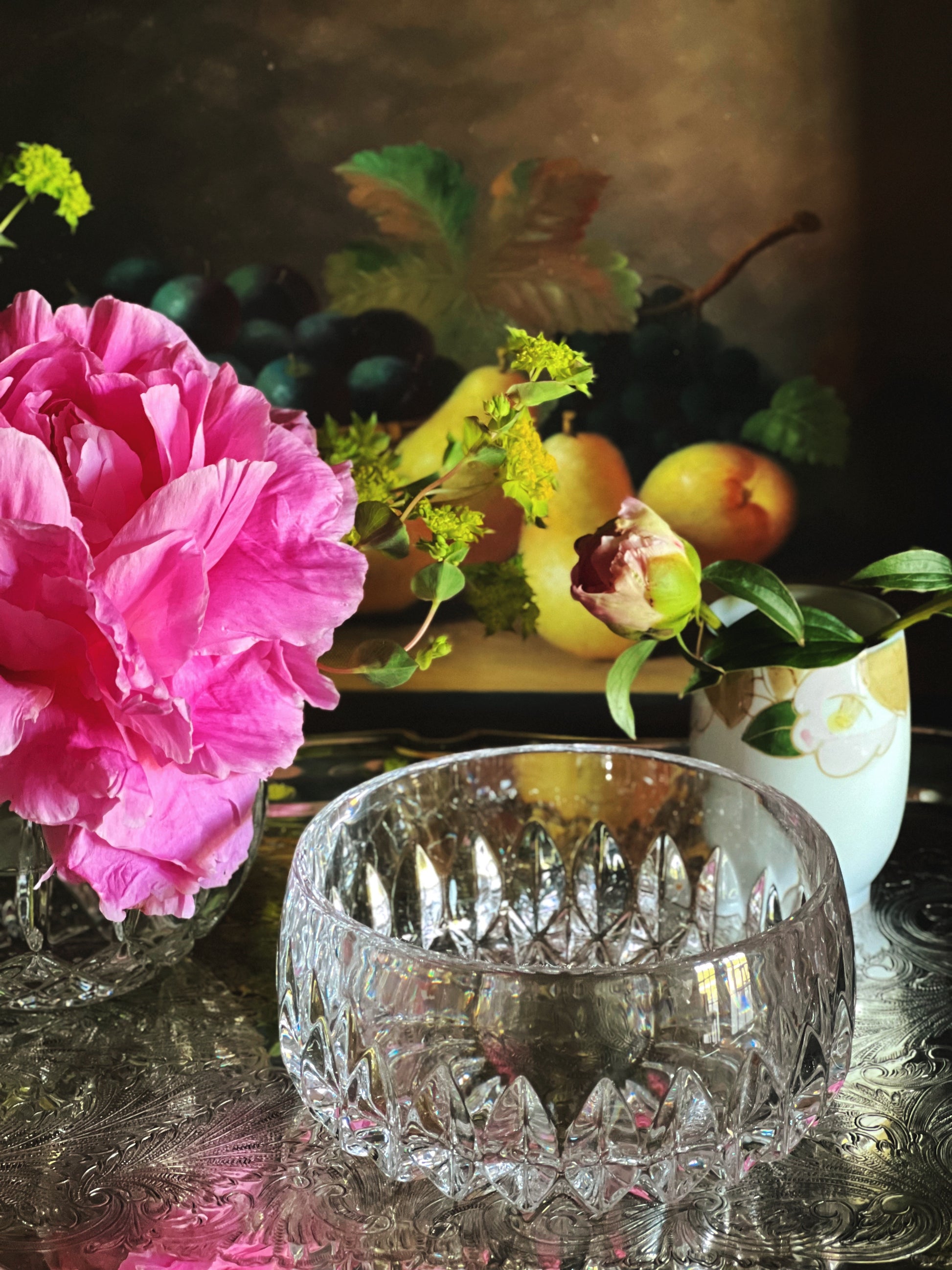 Still life arrangement with pink flowers, a crystal bowl, and fruits on a reflective surface.