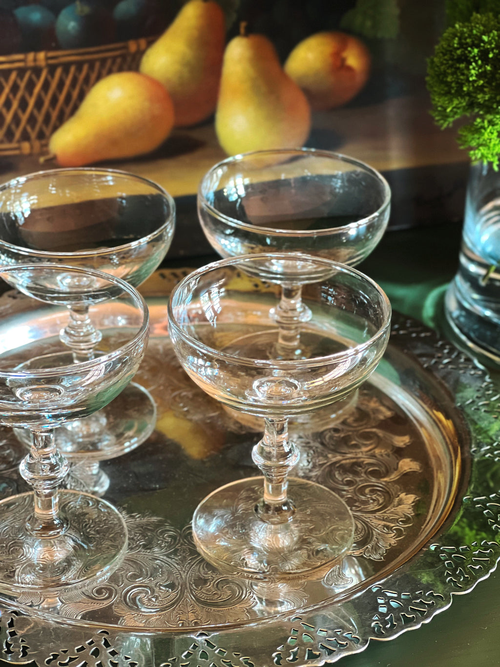 Close up of a set of four vintage champagne coupes with bamboo-style stems displayed on a silver tray with pears and greenery