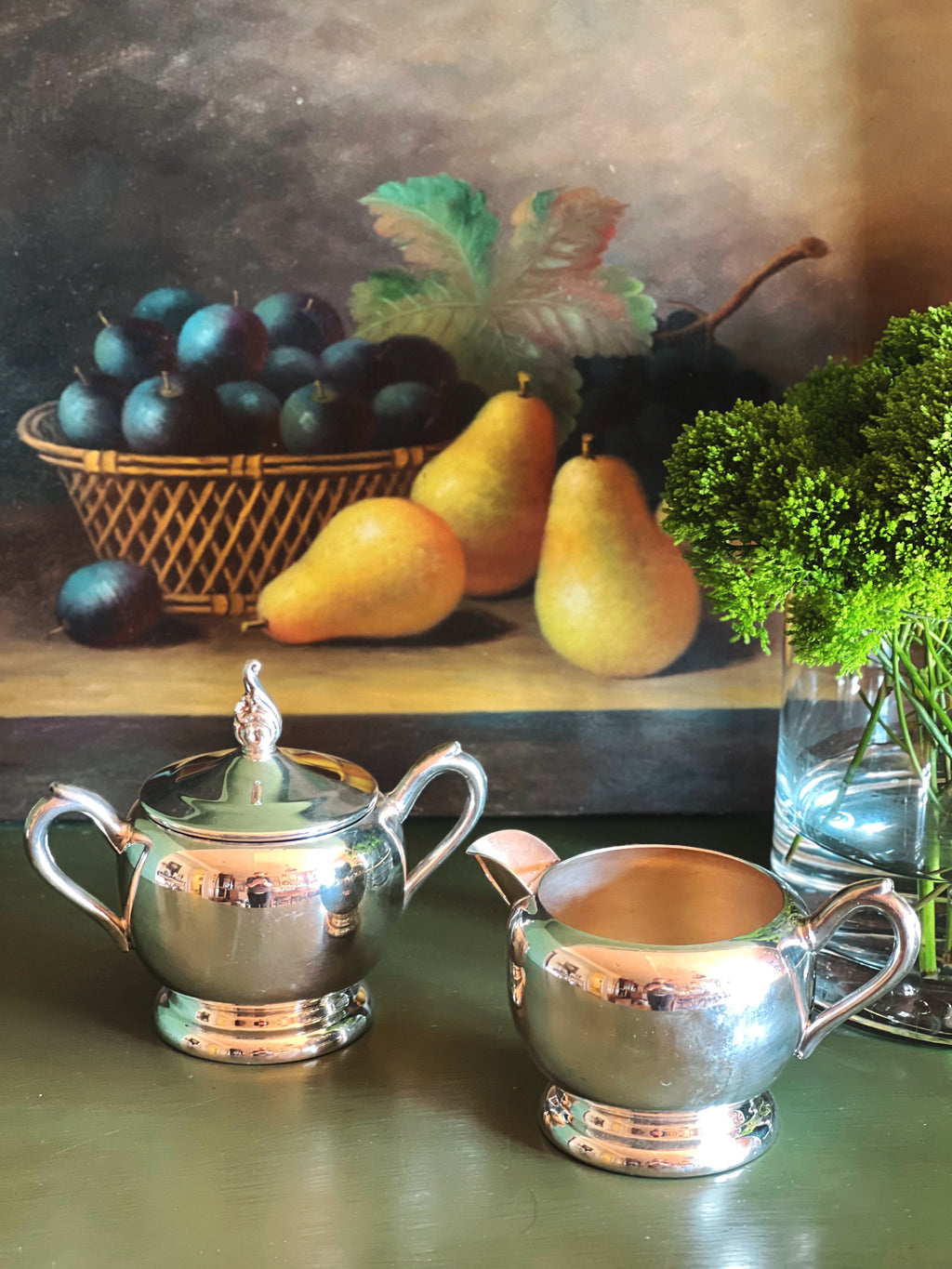 Silver sugar bowl and creamer set on a table with a still life painting in the background.