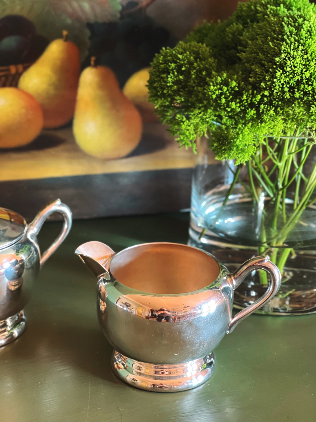 Silver FB Rogers creamer on a table with pears and a vase of greenery in the background