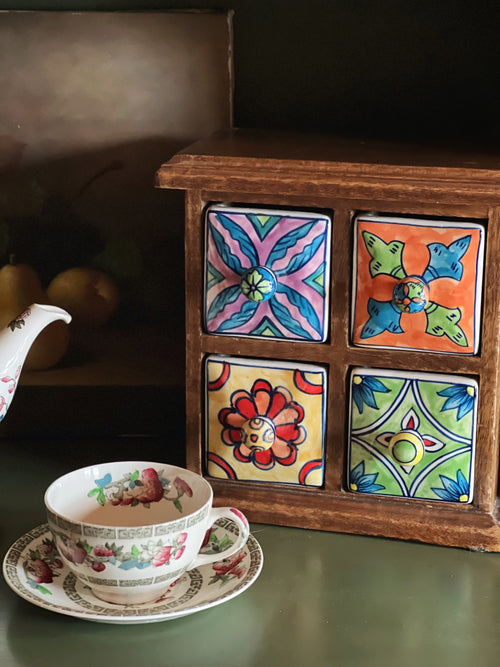 Vintage wooden cabinet with colorful hand painted ceramic drawers styled beside a teacup on a dark green surface